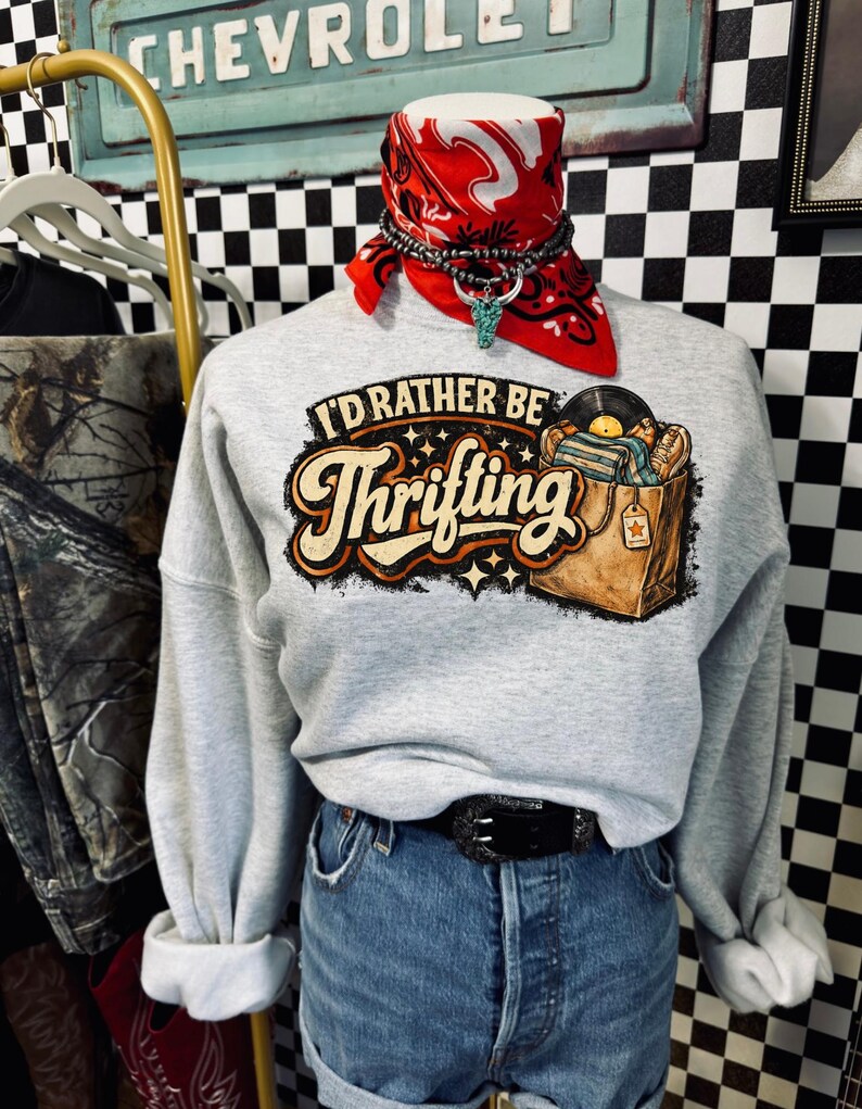 Peut inclure: Un sweat-shirt gris clair avec le texte "I'd Rather Be Thrifting" dans une police r&eacute;tro, avec un graphisme d'un sac de shopping rempli de disques. Le sweat-shirt est associ&eacute; &agrave; un short en jean et une ceinture noire.