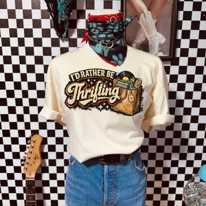 Peut inclure: T-shirt cr&egrave;me avec l'inscription "I'd Rather Be Thrifting" et un graphisme de disques dans une bo&icirc;te. Le t-shirt est associ&eacute; &agrave; un short en jean et une ceinture noire. Un bandana rouge et un collier turquoise ornent le cou du mannequin.