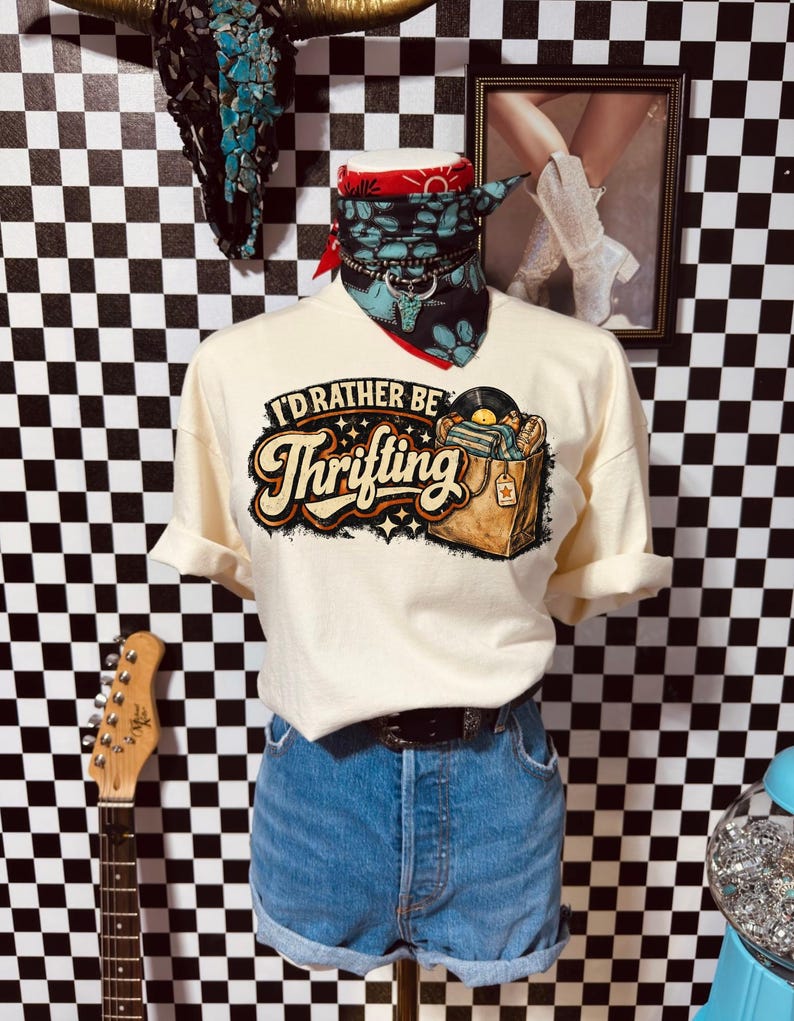 Peut inclure: T-shirt cr&egrave;me avec l'inscription "I'd Rather Be Thrifting" et un graphisme de disques dans une bo&icirc;te. Le t-shirt est associ&eacute; &agrave; un short en jean et une ceinture noire. Un bandana rouge et un collier turquoise ornent le cou du mannequin.