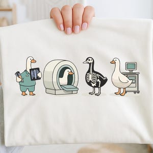 Könnte beinhalten: Ein weißes T-Shirt mit einer Cartoon-Illustration von Gänsen in einer medizinischen Umgebung. Eine Gans hält ein Röntgenbild, eine andere befindet sich in einem MRT-Gerät, eine dritte ist ein Skelett und die letzte steht in der Nähe eines Computers.