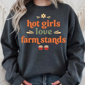 Op de afbeelding: Donkergrijze sweatshirt met de tekst "hot girls love farm stands" in oranje en groen. Het ontwerp bevat een bloem en twee illustraties van jampotten. De sweatshirt wordt gedragen door een persoon die een blauwe jeans draagt.
