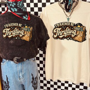 Peut inclure: Deux t-shirts avec le texte "I'd Rather Be Thrifting" et un graphisme d'un tourne-disque et d'un sac de shopping. Un t-shirt est gris fonc&eacute;, l'autre est de couleur cr&egrave;me. Les t-shirts sont expos&eacute;s sur des mannequins.