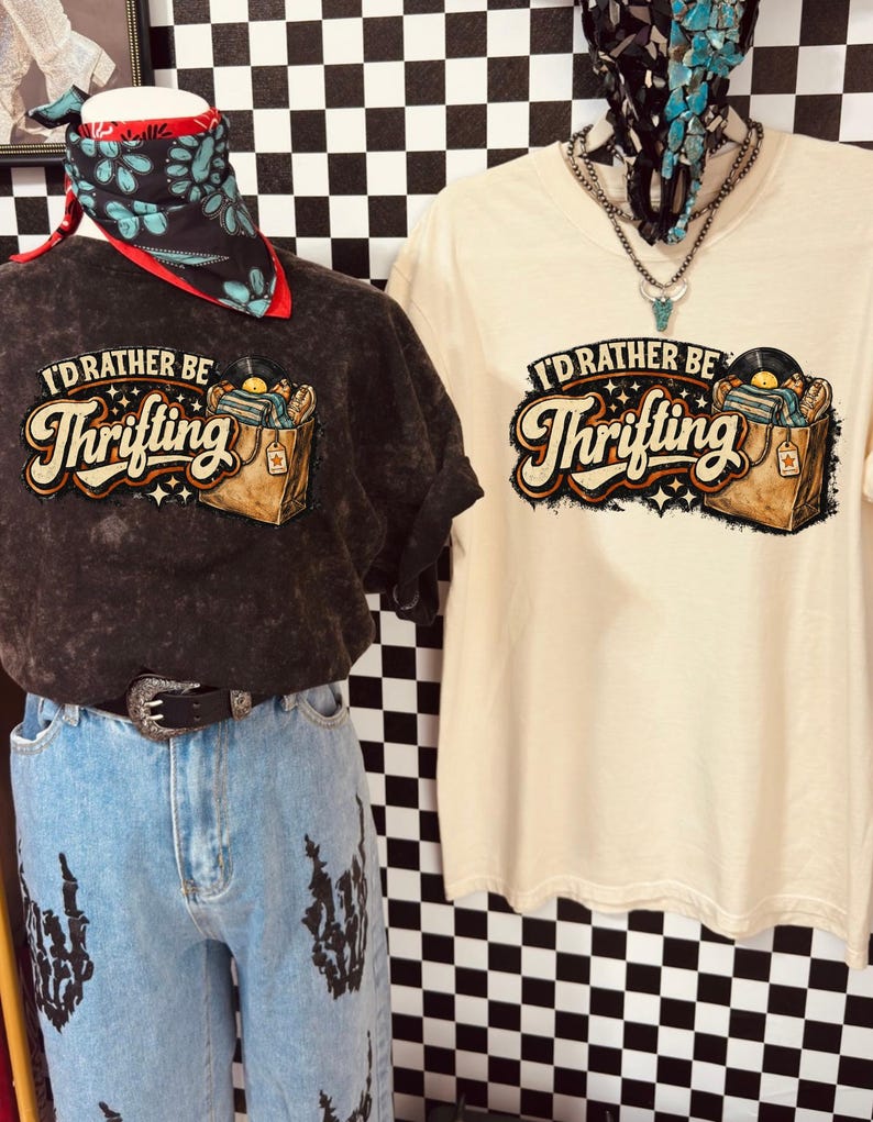 Peut inclure: Deux t-shirts avec le texte "I'd Rather Be Thrifting" et un graphisme d'un tourne-disque et d'un sac de shopping. Un t-shirt est gris fonc&eacute;, l'autre est de couleur cr&egrave;me. Les t-shirts sont expos&eacute;s sur des mannequins.
