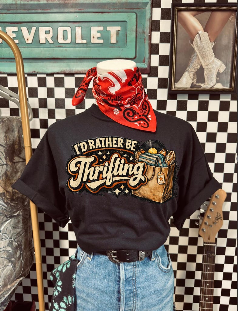 Peut inclure: T-shirt noir avec l'inscription "I'd Rather Be Thrifting" et un graphisme d'une bo&icirc;te remplie de disques vinyles. Le t-shirt est associ&eacute; &agrave; un jean bleu, un bandana rouge et une ceinture noire. Une guitare et une enseigne vintage sont en arri&egrave;re-plan.