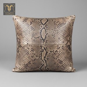 Może przedstawiać: Kwadratowa poduszka dekoracyjna ze wzorem skóry węża w odcieniach beżu, brązu i czerni. Poduszka ma teksturowaną powierzchnię i wzór szwów. Logo Aries Leathers znajduje się w lewym górnym rogu.