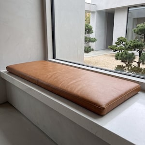 May include: A rectangular, tan-colored leather bench cushion with a smooth, matte finish and clean, straight edges. The cushion is positioned horizontally on a flat, white surface, featuring subtle natural grain textures across its surface.