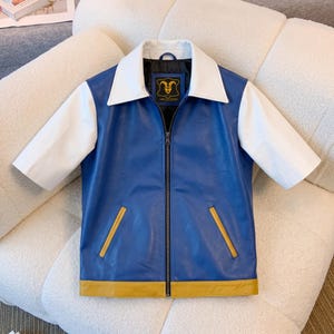 Könnte beinhalten: Eine kurzärmelige Jacke mit blauem Rumpf, weißen Ärmeln und weißem Kragen. Die Jacke hat einen durchgehenden Frontreißverschluss, zwei gelb eingefasste Seitentaschen und einen gelben Saum. Innen befindet sich ein schwarzes Etikett mit Widder-Logo.