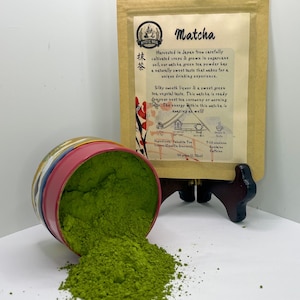 May include: A spilled container of vibrant green matcha powder next to a package of matcha tea. The package has text that reads "Matcha" and describes the tea's origin and flavor profile. The container is a colorful tin.