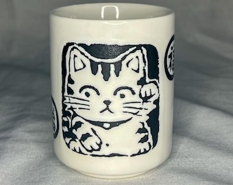 May include: A white ceramic tumbler featuring a black illustration of a Maneki-neko cat with a raised paw. The tumbler has a cylindrical shape and is decorated with Japanese characters. The cat design is centered on the tumbler.