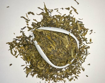 Puede incluir: Hojas de té verde sueltas en un cuenco de cerámica blanco. Las hojas de té están esparcidas alrededor del cuenco sobre un fondo blanco.