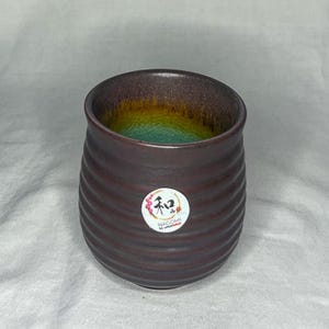 May include: A brown ceramic tumbler with a ridged exterior. The interior features a gradient of colors, including green, yellow, and brown. A white sticker with Japanese text and the word "NAGOMI" is on the side.