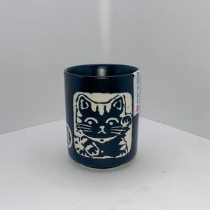 May include: A white ceramic mug with a black handle and the text "But First, Coffee" in a handwritten style. The mug is on a white surface. The mug is approximately 10 cm tall.