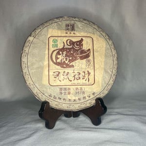 Könnte beinhalten: Ein runder, brauner Teekuchen, verpackt in weißes Papier. Die Verpackung zeigt eine braune Illustration einer Ratte, die einen Goldbarren hält. Der Text auf der Verpackung lautet "Ling Shu Zhao Cai" und "Pu Er Tea (Reifer Tee)".