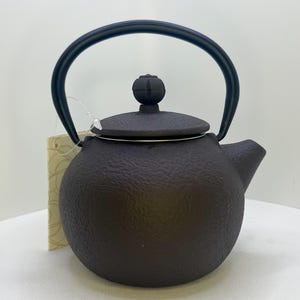 May include: A dark brown, round cast iron teapot with a textured surface, a matching lid featuring a small knob handle, and a curved black metal swing handle. A small, light-colored paper tag is attached to the side of the teapot.