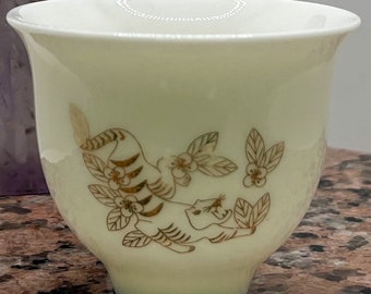 Może przedstawiać: Biały ceramiczny kubek do herbaty z złotym wzorem tygrysa i kwiatów. Kubek stoi na brązowej powierzchni.