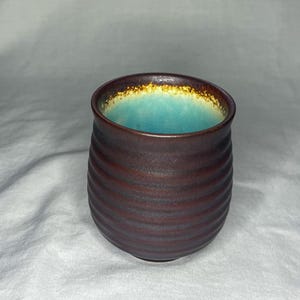 May include: A handmade ceramic tumbler with a dark brown exterior and a turquoise interior. The rim has a gold-colored accent. The exterior features a series of horizontal, ridged lines. The tumbler is designed with a rounded shape.