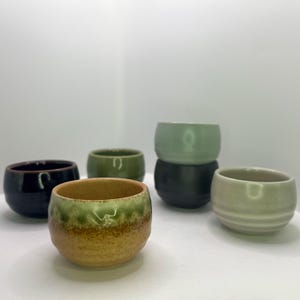 May include: A collection of six small ceramic bowls in various colors, including dark blue, green, and tan. The bowls have a rounded shape and a glazed finish, perfect for serving small portions.