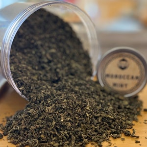 Könnte beinhalten: Getrocknete grüne Teeblätter, die aus einem transparenten Kunststoffbehälter fallen. Eine kleine Dose mit der Aufschrift "MOROCCAN MINT" ist zu sehen. Die Teeblätter sind grün und braun.