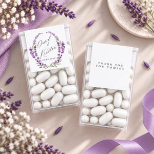 Può includere: Due contenitori rettangolari trasparenti riempiti con caramelle ovali bianche. Uno presenta un'etichetta con una ghirlanda di lavanda ad acquerello, i nomi David & Kristen e la data 26.08.2030. L'altra etichetta riporta THANK YOU FOR COMING.