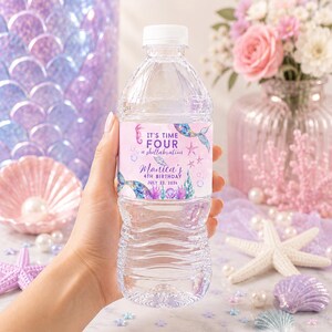Puede incluir: Una botella de agua de plástico transparente con una etiqueta personalizada de temática de sirena, en tonos morados y azules, con el texto: IT'S TIME FOUR a shellabration, Monica's 4TH BIRTHDAY, JULY 23, 2026, decorada con una cola de sirena y coral.