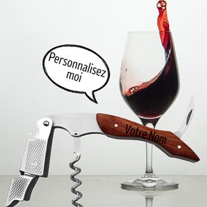 May include: A silver and wood wine opener with a corkscrew and a bottle opener. The opener has a wooden handle with the text "Votre Nom" engraved on it. A glass of red wine is in the background.