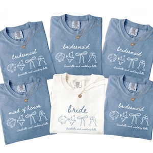 Könnte beinhalten: Eine Auswahl an blauen und weißen T-Shirts mit goldenen Halsketten. Die blauen Shirts tragen die Aufschrift "bridesmaid" oder "maid of honor" mit Muschel-, Bikini-, Schleifen- und Sektglas-Grafiken. Das weiße Shirt trägt die Aufschrift "bride".