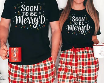 Op de afbeelding: Twee bijpassende zwarte T-shirts met de tekst "SOON TO BE MERRY'D" en kleurrijke lichtslingers. De shirts worden gecombineerd met een rode geruite pyjamabroek. Eén persoon houdt een rode mok met "Merry & Bright", de ander een witte mok.