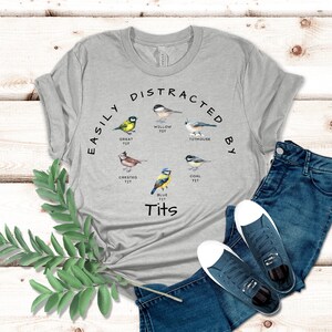 May include: A gray t-shirt with the text "Easily Distracted By Tits" and illustrations of different types of birds, including a great tit, crested tit, willow tit, titmouse, coal tit, and blue tit.