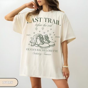 May include: Ivory-colored t-shirt with the text "LAST TRAIL before the veil" and "OLVIA'S BACHELORETTE Gatlinburg, Tennessee" in green. The design includes a pair of boots, a veil, hearts, and the year 2026. The shirt is a casual, oversized fit.