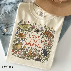 Peut inclure: T-shirt ivoire avec une illustration colorée d'insectes, d'escargots et d'une grenouille, entourés de fleurs et de feuillage. Le texte "LOVE THE UNLOVED" est imprimé en rouge. Le t-shirt est plié sur un jean bleu.