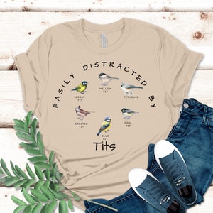 May include: A beige t-shirt with the text "Easily Distracted By Tits" and illustrations of different types of birds, including a blue tit, a great tit, a willow tit, a crested tit, a coal tit, and a titmouse.