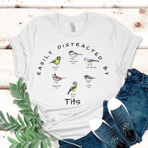 May include: White t-shirt with a black graphic that says "Easily Distracted By Tits" and features illustrations of different types of birds, including a willow tit, great tit, crested tit, blue tit, coal tit, and a titmouse.