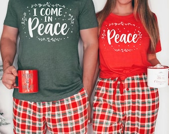 Puede incluir: Conjunto de pijama navideño a juego. Una persona lleva una camiseta verde con el texto "I COME IN Peace" y la otra una camiseta roja con el texto "Peace". Ambos llevan pantalones de cuadros rojos. Cada persona sostiene una taza roja.
