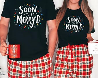 Op de afbeelding: Twee personen dragen bijpassende zwarte t-shirts met de tekst "SOON TO BE MERRY'D" in witte letters, omringd door feestelijke lichtjes en groen. Ze dragen een rode geruite pyjamabroek. Eén persoon houdt een rode mok vast.