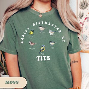 Op de afbeelding: Een groen T-shirt met de tekst "Easily Distracted By Tits" en illustraties van verschillende soorten vogels, die elk met de naam van de vogel zijn gelabeld.