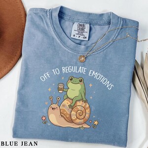 Op de afbeelding: Blauw spijkerbroekkleurig T-shirt met de tekst "OFF TO REGULATE EMOTIONS". De afbeelding toont een groene kikker met een koffiekopje op een slak. Een gouden ketting is over het shirt gedrapeerd.