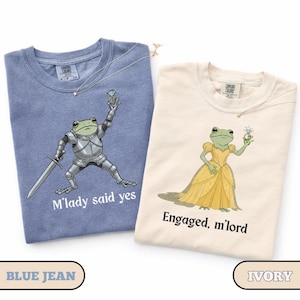 May include: Two t-shirts, one blue and one ivory, with frog illustrations. The blue shirt features a frog in armor with a sword, text reads "M'lady said yes." The ivory shirt shows a frog in a yellow dress, text reads "Engaged, m'lord."