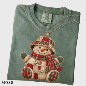 Può includere: Una t-shirt verde muschio con etichetta Comfort Colors e una grafica di un pupazzo di neve patchwork con cappello e sciarpa. Il pupazzo è composto da quadrati di tessuto rosso, verde e crema con due bottoni marroni sul busto.
