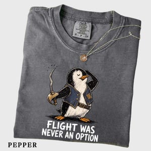 May include: A charcoal gray t-shirt featuring a graphic of a penguin dressed as a pirate, wearing a blue vest and holding a curved sword. The text below the penguin reads, "FLIGHT WAS NEVER AN OPTION." The shirt has a Comfort Colors brand label.