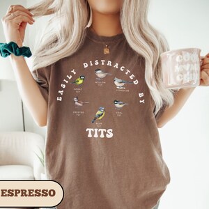 Op de afbeelding: Bruin T-shirt met de tekst "Easily Distracted By Tits" en illustraties van verschillende soorten vogels met hun namen, waaronder "Great Tit", "Willow Tit", "Coal Tit", "Crested Tit" en "Blue Tit".