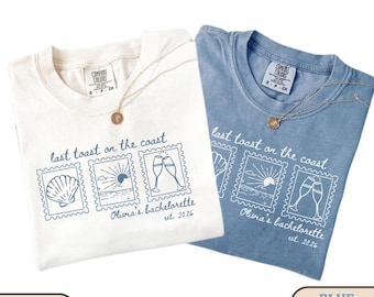 Op de afbeelding: Twee Comfort Colors t-shirts, ivoor en jeansblauw, met de tekst "last toast on the coast" en een postzegelontwerp. Het postzegelontwerp bevat een schelp, zonsondergang en champagneglazen. Elke shirt heeft een gouden ketting.