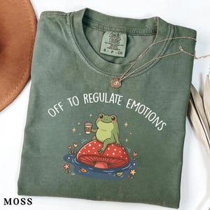 May include: A moss green t-shirt with the phrase "OFF TO REGULATE EMOTIONS" and a cartoon frog holding a coffee cup. The frog is sitting on a red mushroom with white spots. A silver necklace is draped on the shirt.