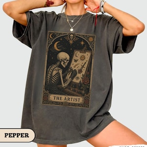 Può includere: Una t-shirt grigio antracite con una stampa grafica rettangolare di uno scheletro che dipinge su un cavalletto. L'opera include motivi celesti come una luna crescente, stelle e un sole, con il testo THE ARTIST in fondo alla cornice.