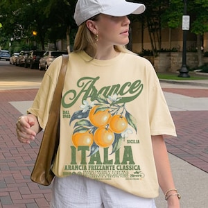 Woman standing outdoors wearing a pale yellow Comfort Colors t-shirt with a vintage Italian orange soda graphic. The design features bold green and orange typography with illustrations of oranges and the words "Arancia Frizzante Classica" and "Italia Sicilia." The model is 5'5" and wearing an XL for an oversized fit. Urban background with trees and brick road.
