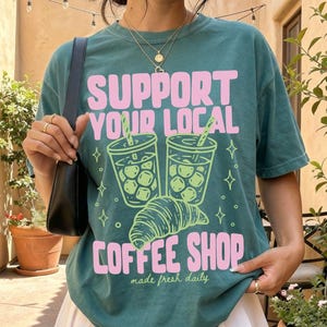 May include: A blue spruce t-shirt featuring a graphic of two iced coffee cups and a croissant with the text SUPPORT YOUR LOCAL COFFEE SHOP and made fresh daily in pink and light green, paired with a white pleated skirt and a black shoulder bag.