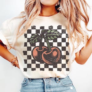 Peut inclure: Un t-shirt blanc avec un motif à carreaux et un graphique de cerise de style vintage. Le texte "COLOR IVORY" est imprimé en bas de l'image.