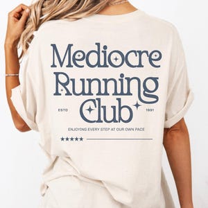May include: An ivory short-sleeve t-shirt featuring dark blue text on the back that reads Mediocre Running Club, ESTD 1991, and ENJOYING EVERY STEP AT OUR OWN PACE, accompanied by small star icons and a line of five stars above a horizontal line.