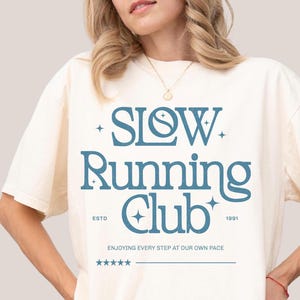 Op de afbeelding: Crèmekleurig T-shirt met de tekst "SLOW Running Club" in teal. Het shirt bevat ook de tekst "ESTD 1991" en "ENJOYING EVERY STEP AT OUR OWN PACE". Het model is 1,73 m lang en draagt XL.