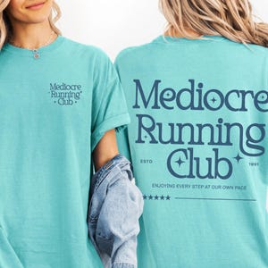 以下が含まれることがあります： ダークブルーのテキストがプリントされたターコイズブルーの半袖Tシャツ。左胸には「Mediocre Running Club」の文字。背面には同じテキストが大きくプリントされ、「ESTD 1991」、「ENJOYING EVERY STEP AT OUR OWN PACE」、5つの星がデザインされています。