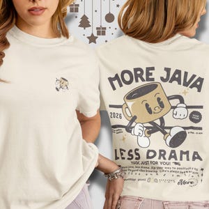 May include: Cream-coloured t-shirt featuring a cartoon coffee cup design on the front and back. The back of the shirt displays the text "MORE JAVA LESS DRAMA" with a coffee cup character and the year "2026". The t-shirt has short sleeves.